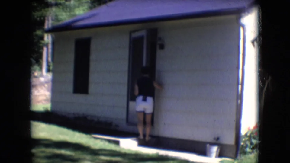 1966 Athens, Ohio home-movie frame with a woman seen from behind at the open door of a residential house, with lawn and trees around it.