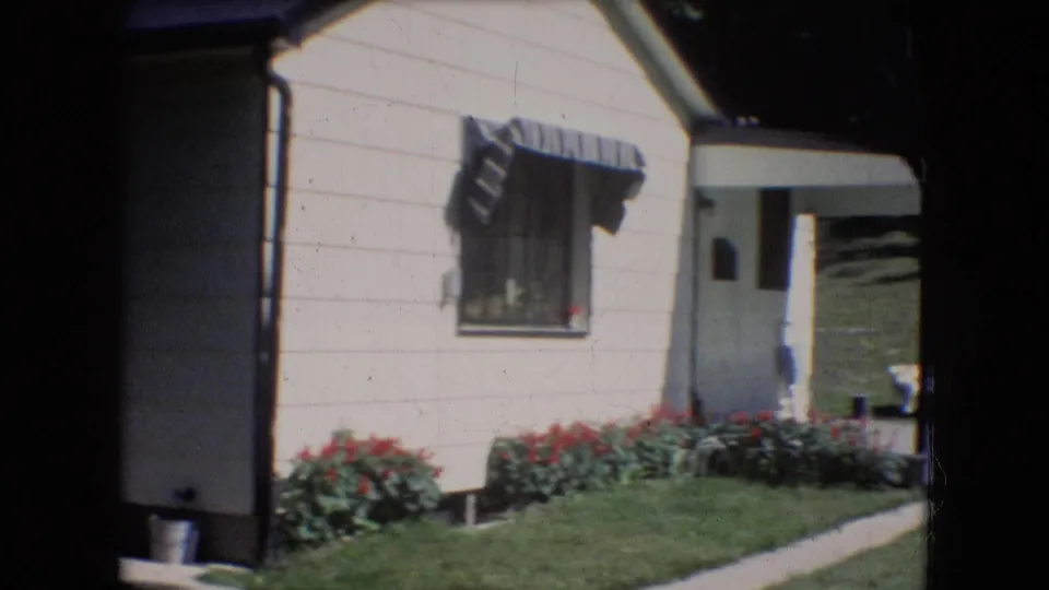 1966 Athens, Ohio 8mm frame showing the side of a light-colored house with a window awning, flower bed, lawn, walkway, and shaded entry area.