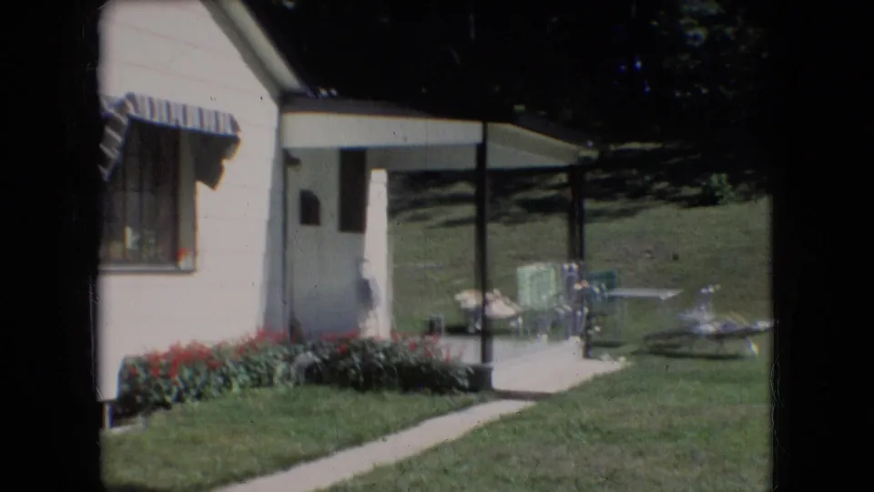 1966 Athens, Ohio home-movie frame showing a white-sided house with a striped awning, red flowers, green lawn, concrete walkway, and outdoor chairs.
