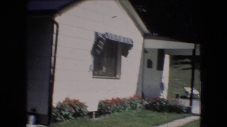 1966 Athens, Ohio frame of the house exterior with a striped window awning, red flowers along the wall, lawn, and a shaded doorway area.