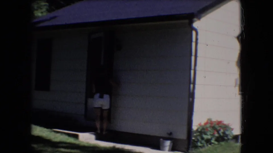 1966 Athens, Ohio home-movie frame showing a woman in a dark top and light shorts near the doorway of a white-sided house, with flowers and lawn visible nearby.