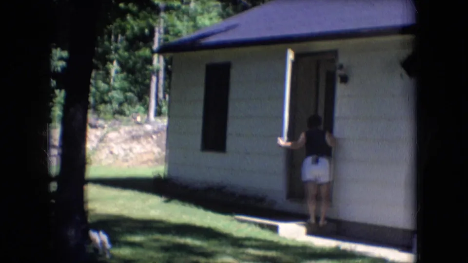 1966 Athens, Ohio frame showing a woman at the open front door of a white-sided house, with a shaded lawn and trees in the background.
