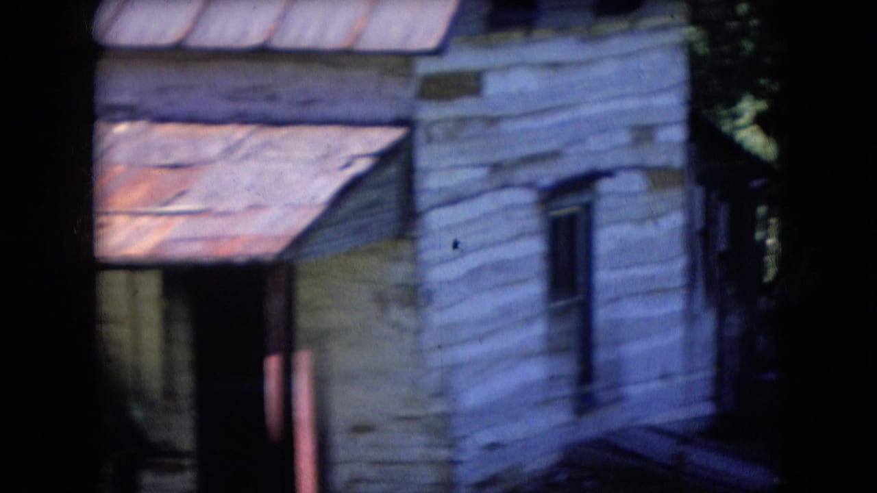 1966 Athens, Ohio 8mm frame showing a weathered white-sided cabin or outbuilding with a rusted metal roof, dark window, and surrounding trees.