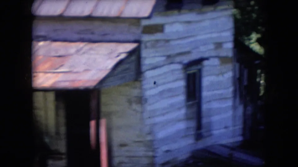 1966 Athens, Ohio 8mm frame showing a weathered white-sided cabin or outbuilding with a rusted metal roof, dark window, and surrounding trees.