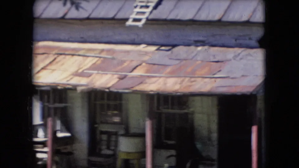 1966 Athens, Ohio 8mm frame showing the front of a weathered cabin with a rusted metal porch roof, posts, windows, and a dark open doorway.