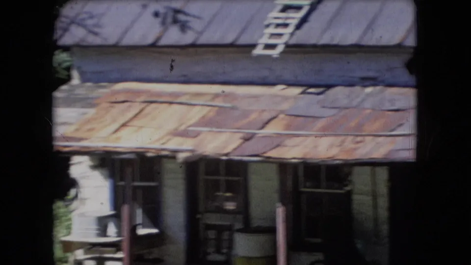 1966 Athens, Ohio 8mm frame showing a weathered cabin front with rusted metal roofing, a ladder near the roofline, windows, porch posts, and a dark doorway where a person may be partially visible.