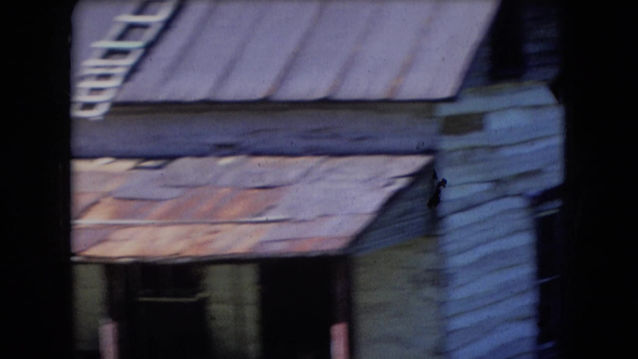 Athens, Ohio 1966 8mm frame showing a close view of a weathered white house or cabin with rusted metal roofing and shaded porch details.