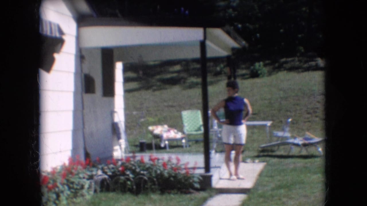 Woman in a blue top and white shorts standing on a porch near a lawn and flowers.