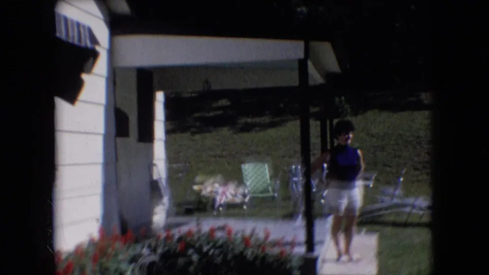 Athens, Ohio 1966 8mm frame showing a woman standing under a porch roof near a post, with vintage-style lawn chairs, flowers, and a shaded grassy yard visible behind her.