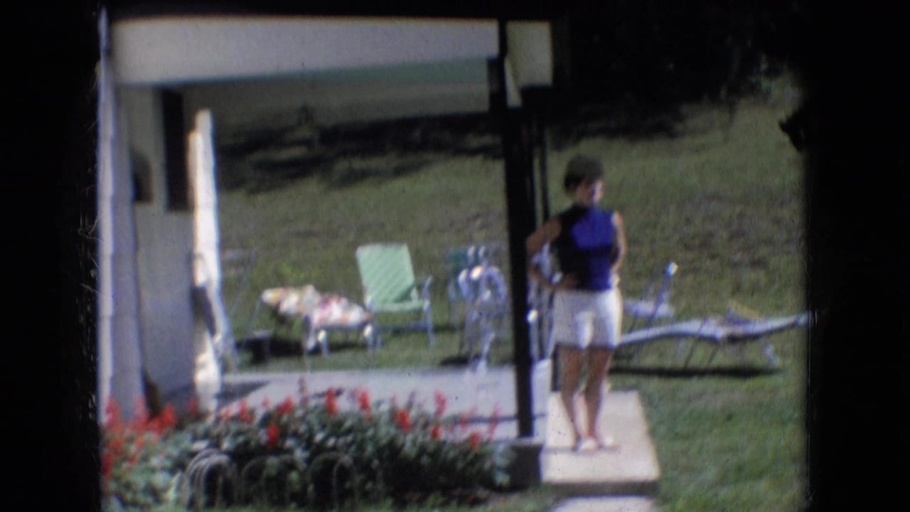 In Athens, Ohio, 1966 home-movie footage, a woman stands near a porch in a blue sleeveless top and white shorts with lawn chairs, grass, and red flowers visible around her.