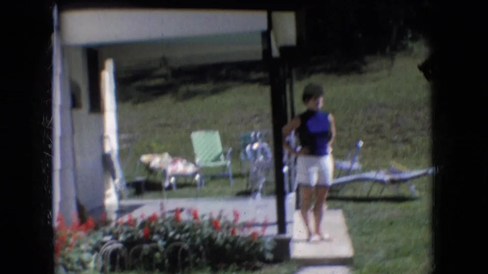In Athens, Ohio, 1966 home-movie footage, a woman stands near a porch in a blue sleeveless top and white shorts with lawn chairs, grass, and red flowers visible around her.