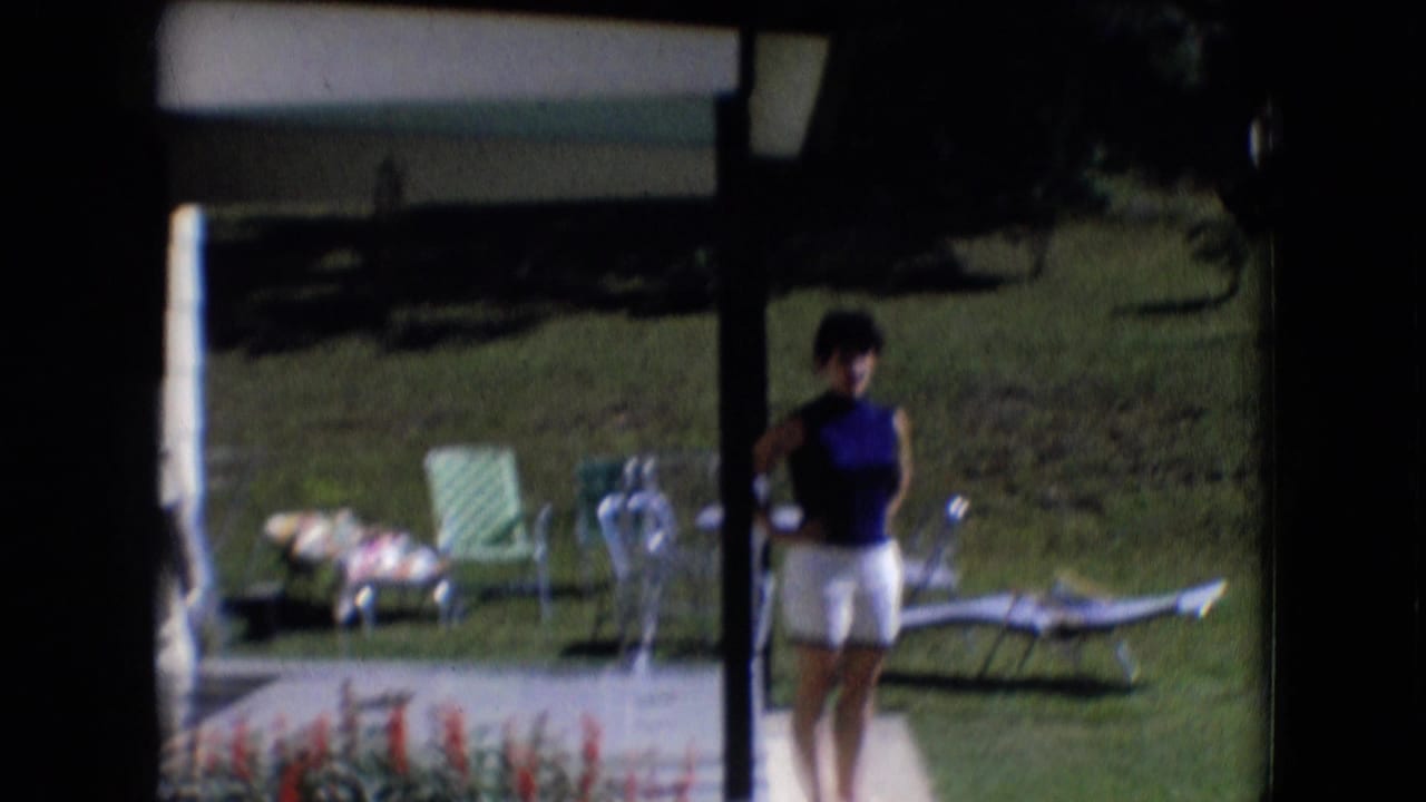 The woman remains on the porch or walkway in the 1966 Athens, Ohio home-movie frame, facing the camera with green chairs and a lounge chair set on the lawn behind her.