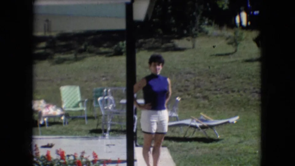 A woman in a blue top and white shorts stands with hands on hips near a porch post in Athens, Ohio, with red flowers in the foreground and lawn furniture in the background.