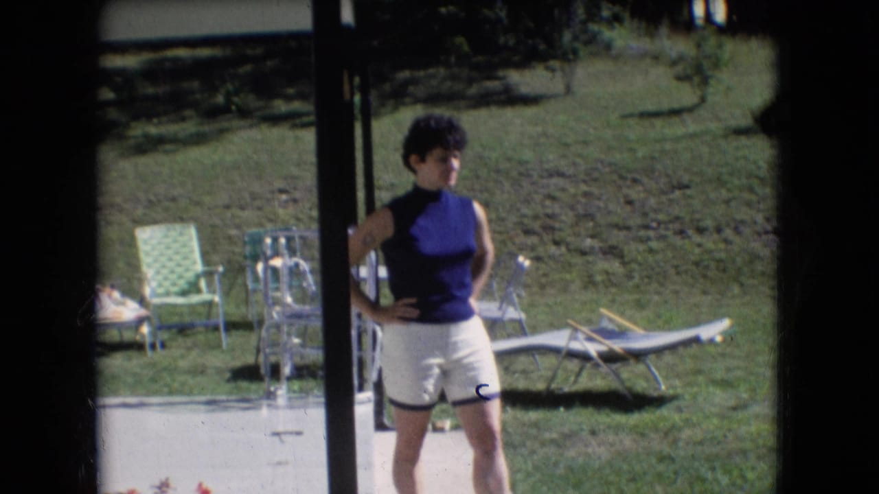 In Athens, Ohio home-movie footage from 1966, a woman in a sleeveless blue top and white shorts stands near a patio with lawn chairs and a grassy yard behind her.
