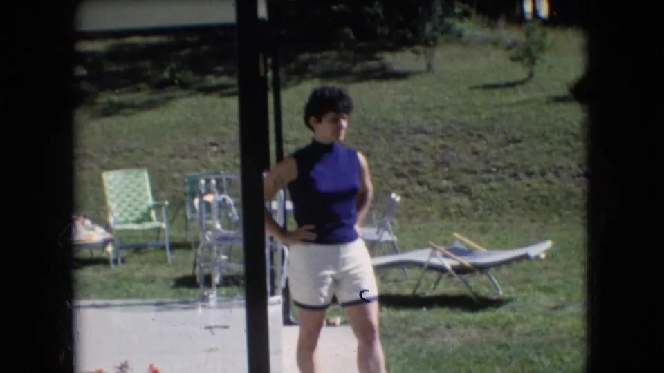 In Athens, Ohio home-movie footage from 1966, a woman in a sleeveless blue top and white shorts stands near a patio with lawn chairs and a grassy yard behind her.