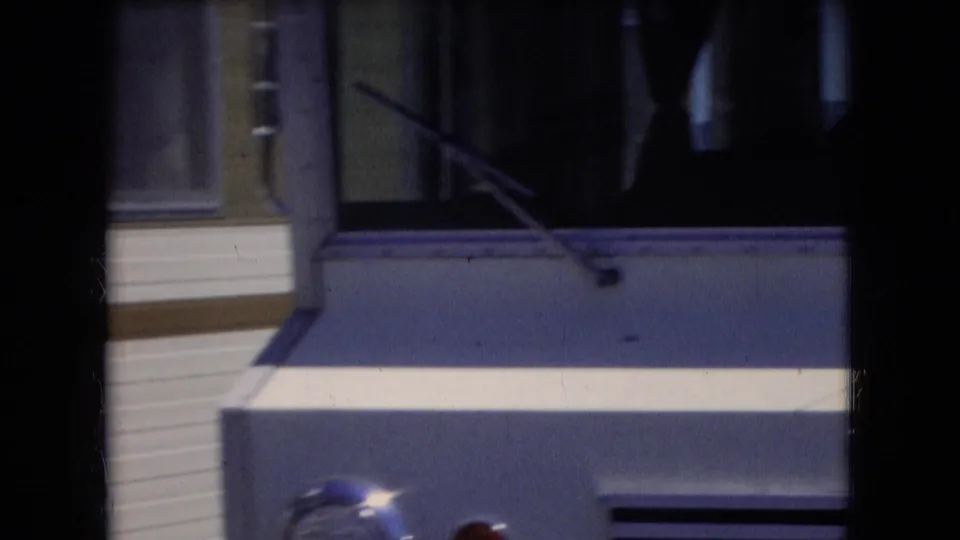 Athens, Ohio 1966 home-movie frame continuing across the white-and-blue RV, showing windows, body panels, dark interior reflections, and front trim.