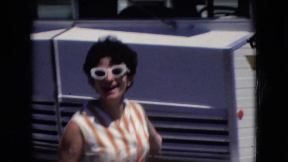In supplied 1966 Athens, Ohio home-movie footage, the woman smiles toward the camera while standing in front of the mobile-home exterior.