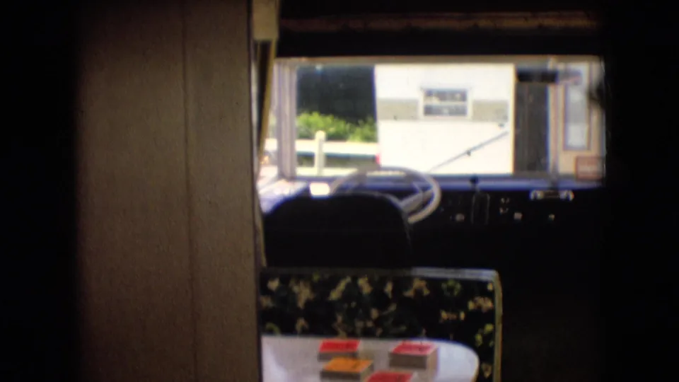 The interior frame shows a vehicle or mobile-home cabin with steering wheel, dashboard, windshield view, floral-patterned table, and small boxed items or cards.