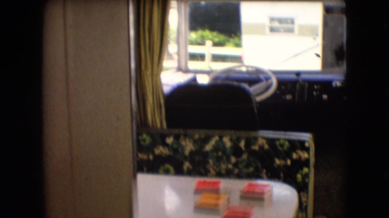 1966 Athens, Ohio 8mm frame showing the front of a camper interior with a large steering wheel, dashboard windows, curtains, patterned seating, and a small table with game pieces.