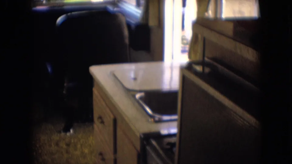 1966 Athens, Ohio 8mm frame showing the camper kitchenette area with a sink cabinet, side window, patterned seat back, front chair, and carpeted floor.