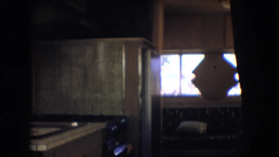 1966 Athens, Ohio 8mm frame of a dim camper interior showing a kitchenette counter, cabinet, window light, dark seating, and part of the driving area.