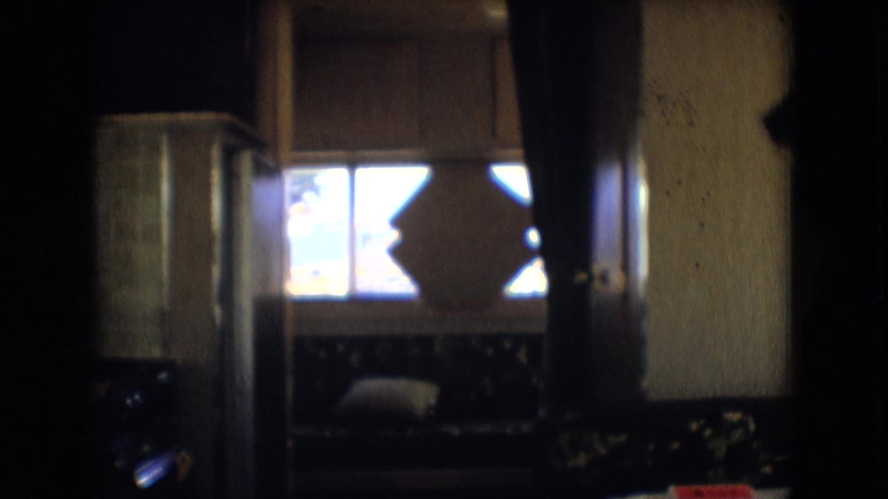 Dim 1966 home-movie view inside a motorhome in Athens, Ohio, showing a window with an X-shaped frame detail and a cushioned bench seat.