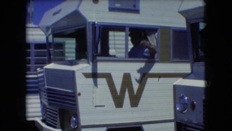 A man leans out from the driver-side window of a white Winnebago motorhome in 1966 Athens, Ohio, with the large side W logo visible below him.