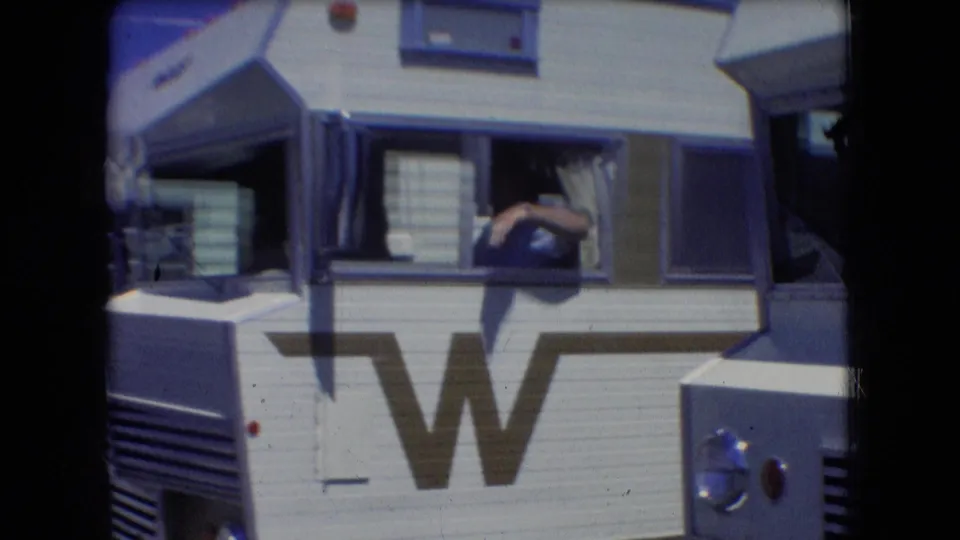 Side view of the white Winnebago motorhome in 1966 Athens, Ohio, with a driver's arm visible through the cab window and the large W logo across the body panel.