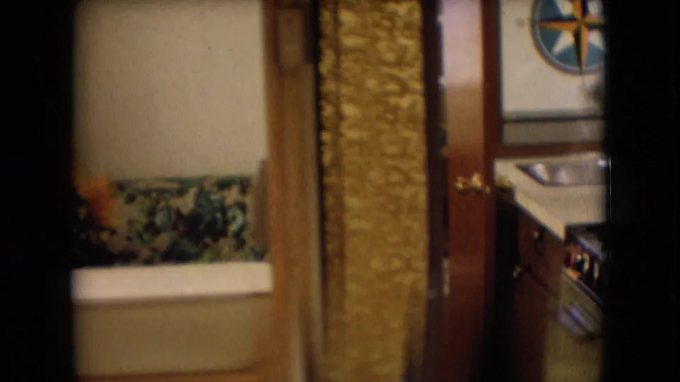Handheld 8mm view through a narrow motorhome interior, with a light-colored bath or seating fixture, patterned fabric, textured wall panel, and partial kitchen doorway.