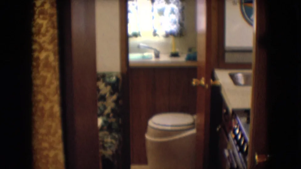 1960s motorhome bathroom view showing a toilet, small window with patterned curtain, wood wall panels, and the adjacent compact kitchen area.