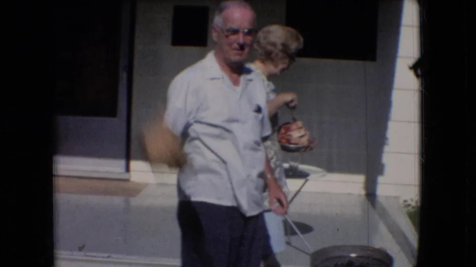 In supplied 1966 Athens, Ohio home-movie footage, an older man in a white short-sleeve shirt stands near a porch as an older woman in glasses turns beside him.