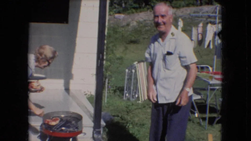 The older man smiles in the sunny Athens, Ohio backyard, with lawn chairs and laundry on a clothesline visible behind him in the 1966 home-movie frame.