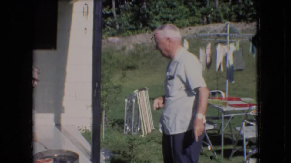 In a 1966 Athens, Ohio backyard scene, an older man in a light shirt stands near porch steps while lawn chairs and a clothesline are visible in the yard.
