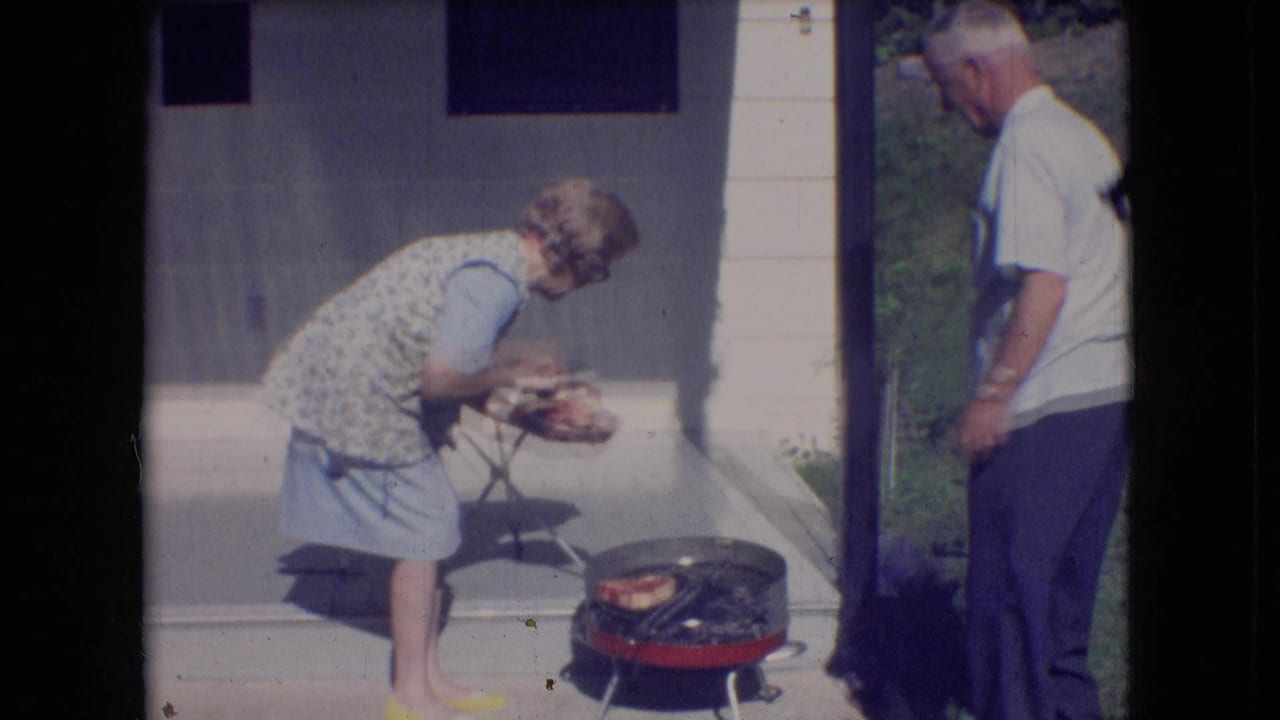 Older woman bends over a small red grill while an older man stands nearby.