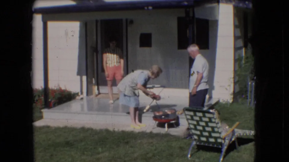 A wider 1966 Athens, Ohio backyard view shows the older couple near the red grill, a lawn chair in the foreground, and a younger person standing in the doorway.
