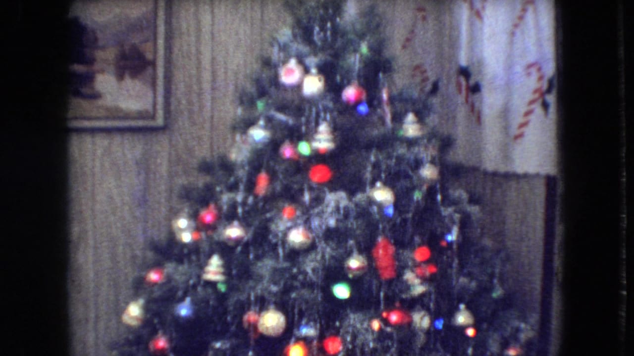 1966 Athens, Ohio home-movie frame showing a decorated Christmas tree with colored lights, ornaments, tinsel, wood-paneled wall, framed picture, and patterned curtains.