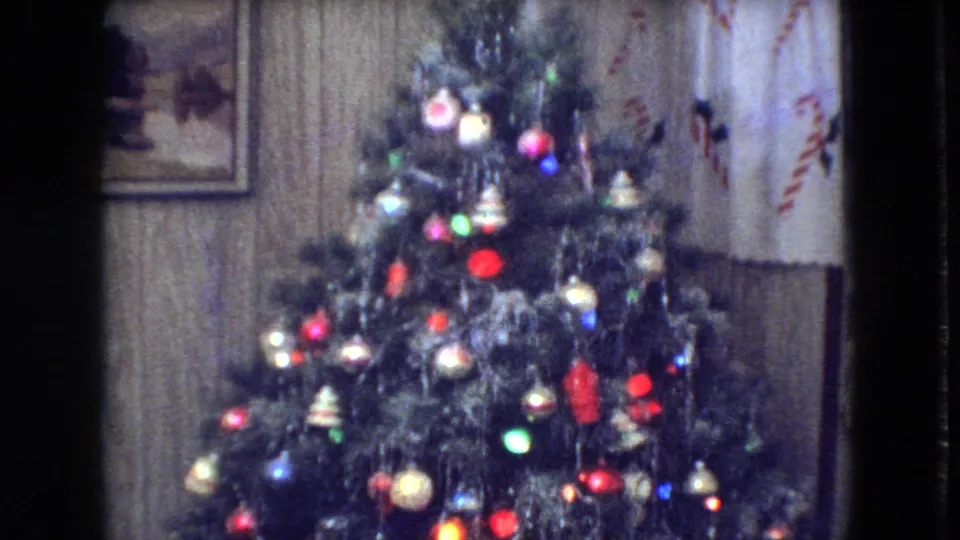 1966 Athens, Ohio home-movie frame showing a decorated Christmas tree with colored lights, ornaments, tinsel, wood-paneled wall, framed picture, and patterned curtains.