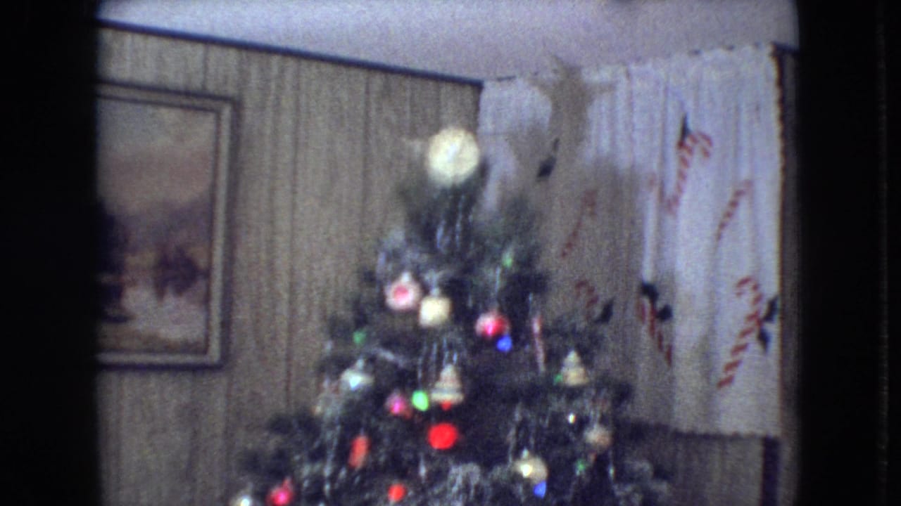 1966 Athens, Ohio home-movie frame showing the upper section of a decorated Christmas tree with a glowing topper, colored ornaments, wood paneling, framed wall art, and candy-cane patterned curtains.