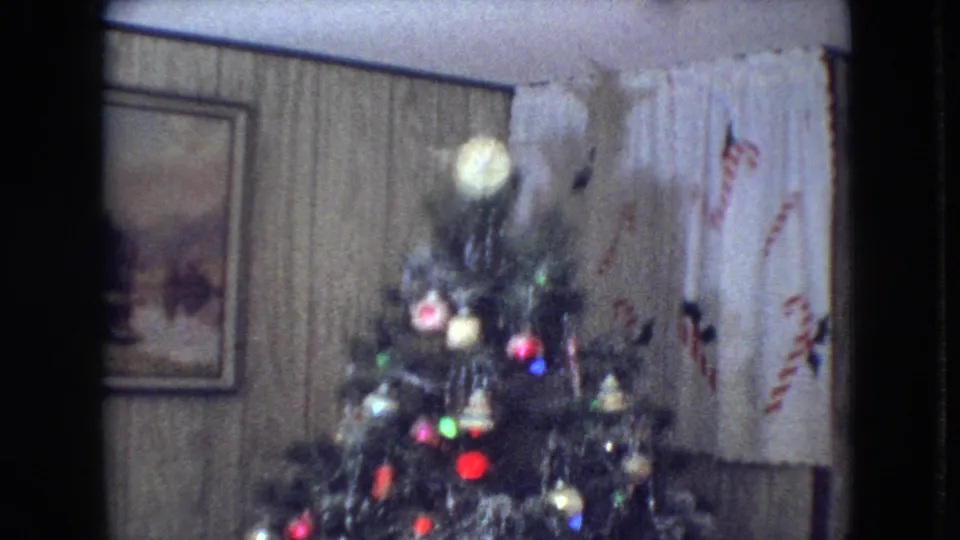 1966 Athens, Ohio home-movie frame showing the upper section of a decorated Christmas tree with a glowing topper, colored ornaments, wood paneling, framed wall art, and candy-cane patterned curtains.