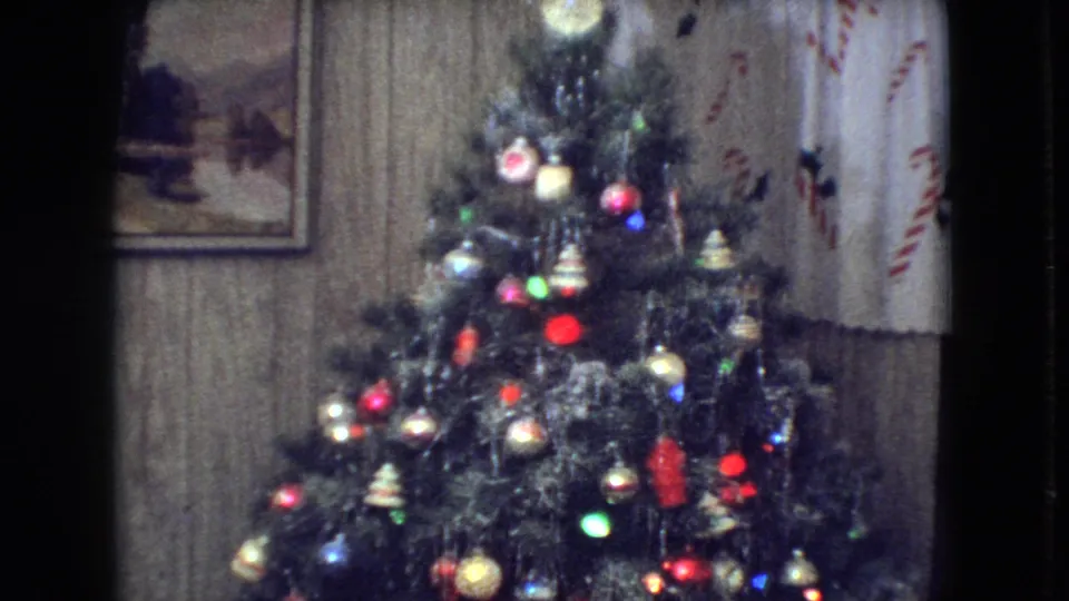 1966 Athens, Ohio home-movie frame showing a decorated Christmas tree filling the frame, with tinsel, multicolored ornaments, lights, wood-paneled walls, framed artwork, and patterned curtains visible behind it.