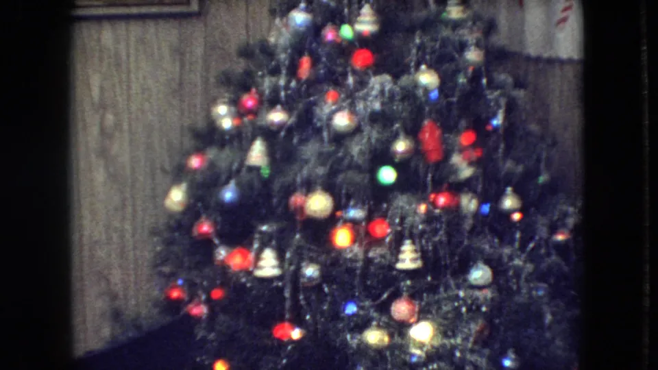 1966 Athens, Ohio home-movie frame showing a fuller view of the Christmas tree with colored lights, ornaments, hanging decorations, tinsel, wood paneling, framed wall art, and patterned curtains.