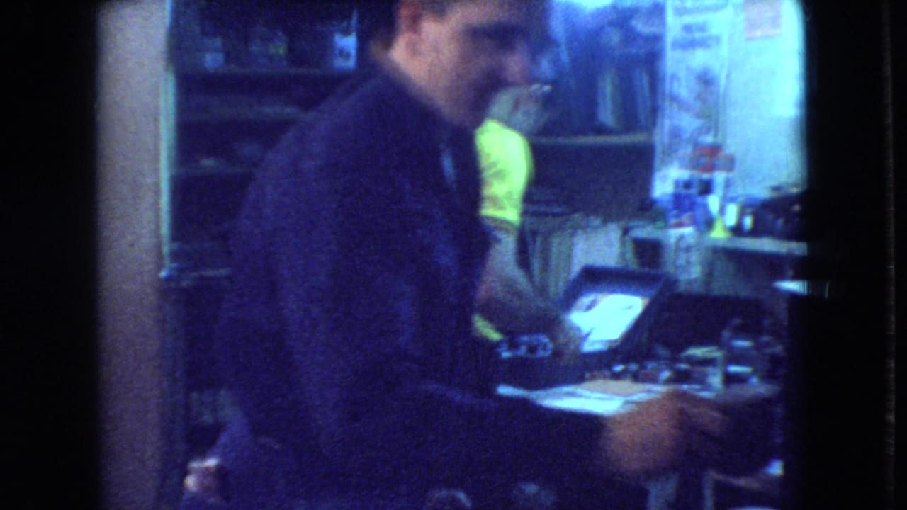 In Athens, Ohio 1966 home-movie footage, a man in dark clothing turns toward the camera and smiles beside a dim workshop table where another man works.