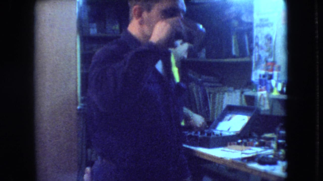 Man in dark clothing drinks from a mug beside a cluttered workshop table.