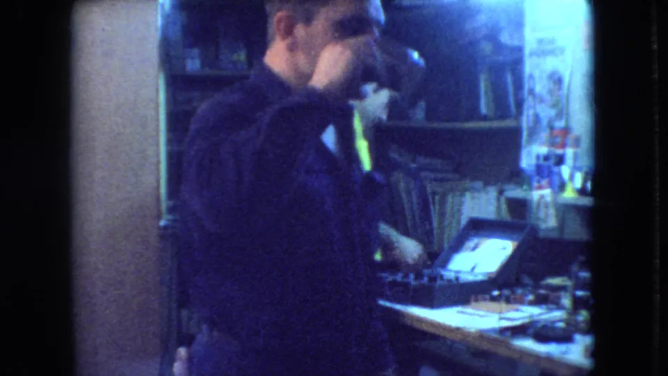 In the dim Athens, Ohio workshop scene, the man raises a mug to drink while another adult remains at the workbench in the background.