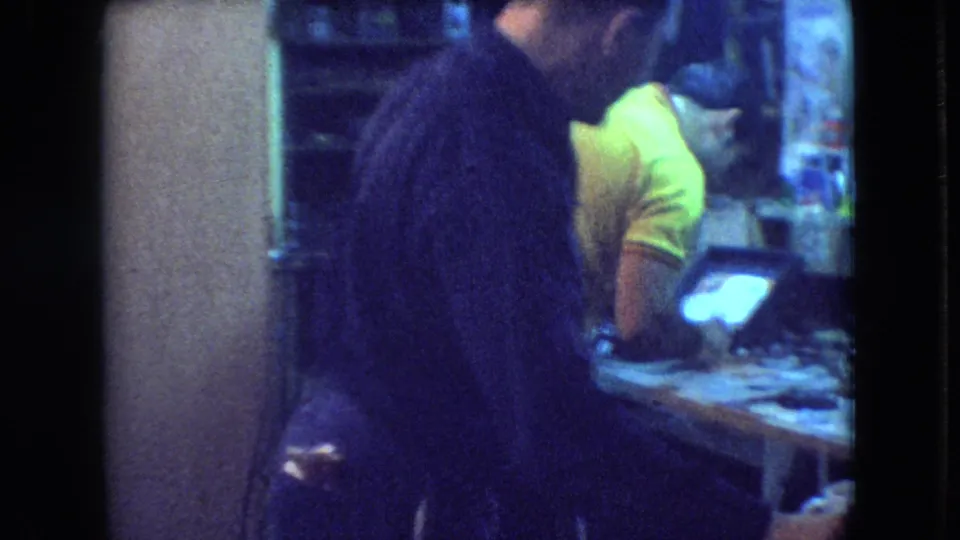 The home-movie frame shows the dark-clothed man in the foreground and a man in a bright yellow shirt bending over a cluttered workshop table.