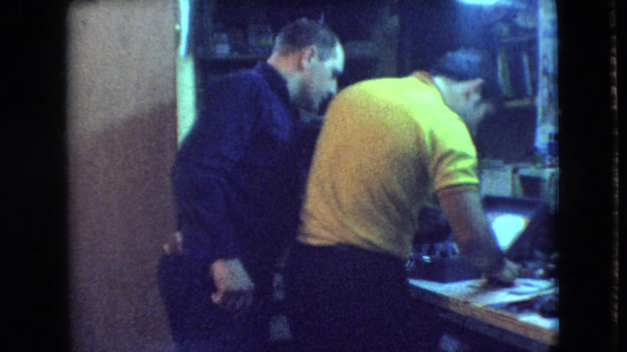 In a 1966 Athens, Ohio home-movie frame, a man in a yellow polo works at a crowded bench while a man in a dark shirt watches beside him.