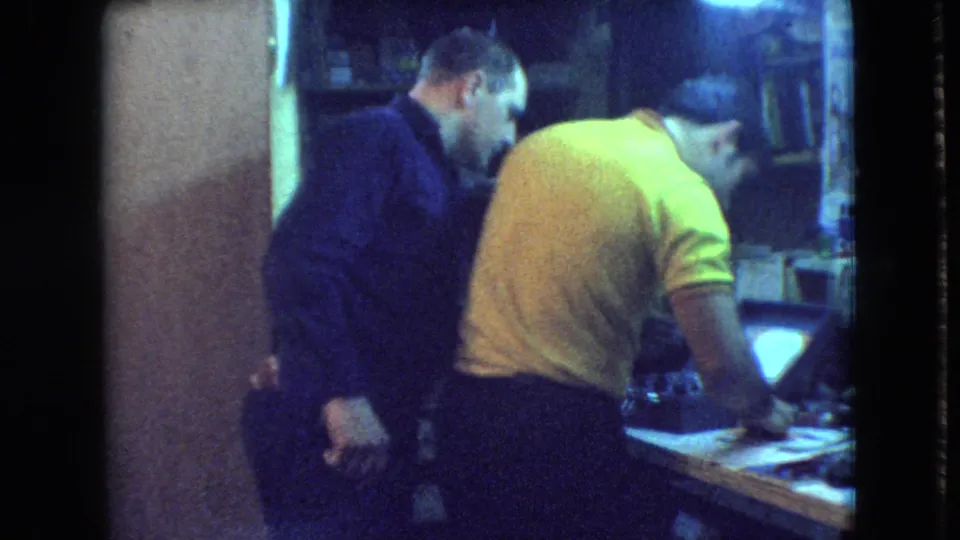 In a 1966 Athens, Ohio home-movie frame, a man in a yellow polo works at a crowded bench while a man in a dark shirt watches beside him.