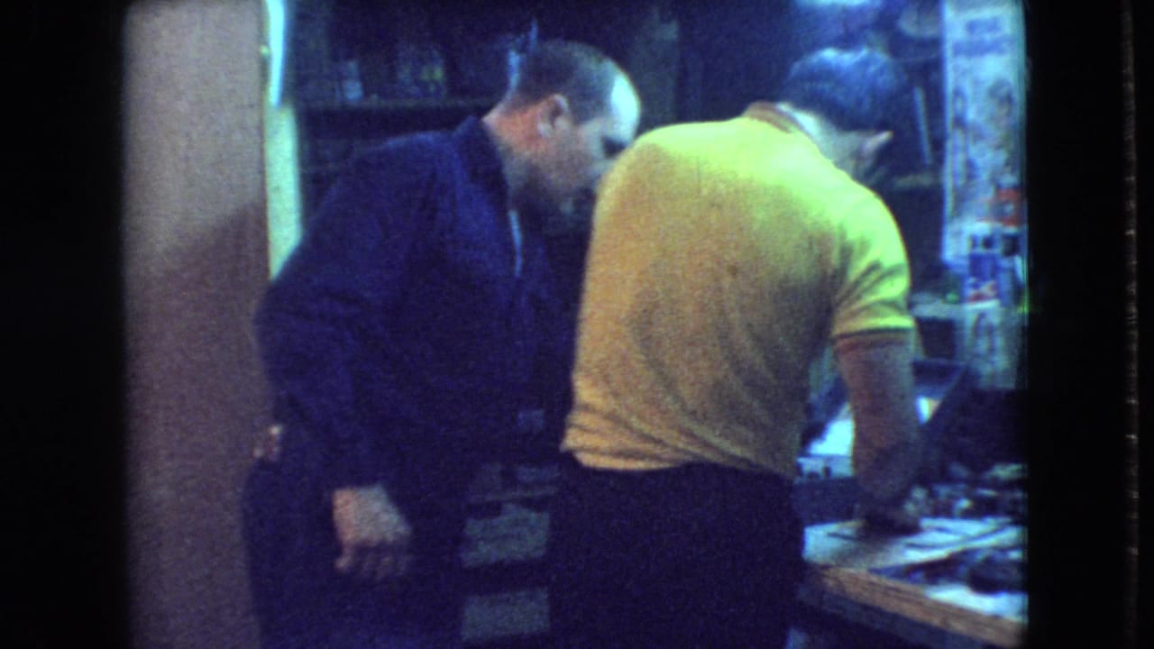 Two men lean over a workbench with tools and parts in a small room.