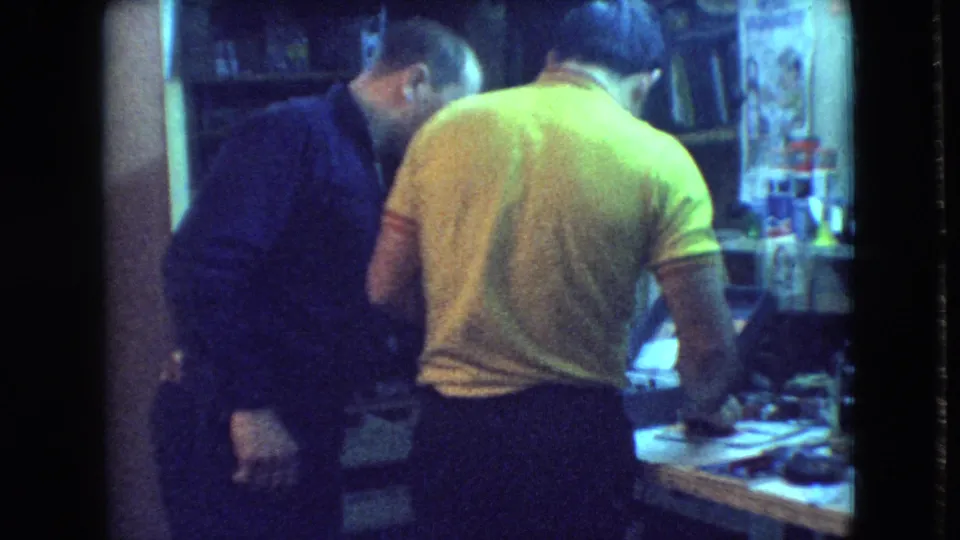 A 1960s home-movie frame shows the man in yellow turned toward the work surface, with another adult man partly behind him in the private workshop.