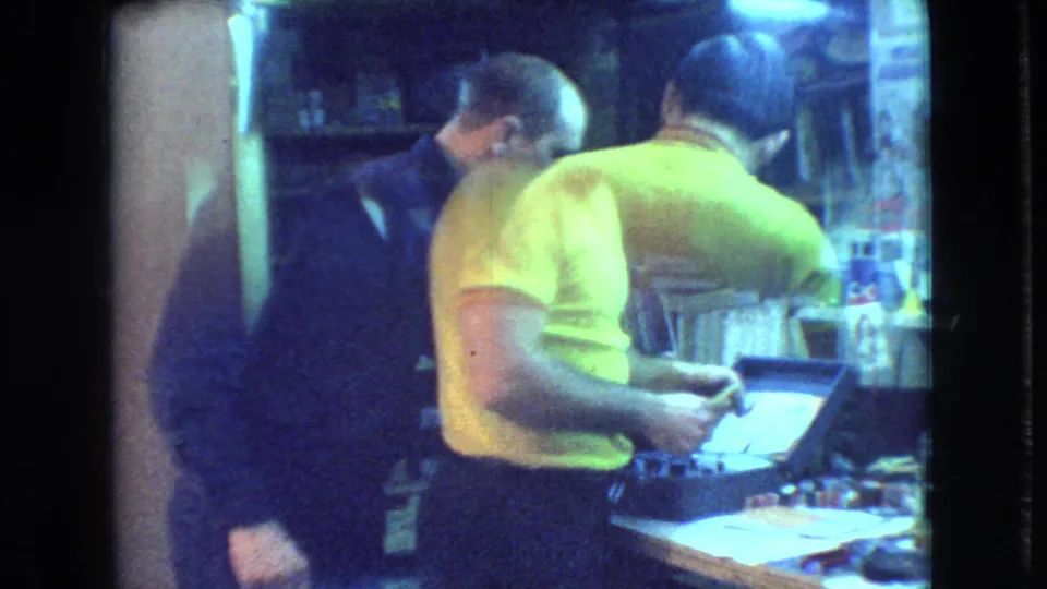 At the end of the Athens, Ohio clip, the man in the yellow polo appears to handle a small object near the bench while the second man remains nearby in the dim room.